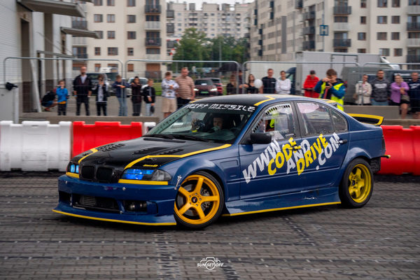 BMW E46 drifting on a city car park, spectators behind the barriers and apartment blocks in the background