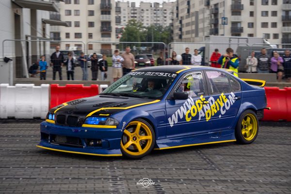 Blue and black BMW E46 drift car with yellow wheels sliding in front of the spectators